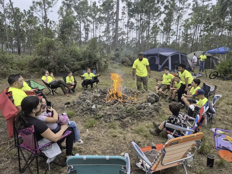 Scouts and families sit in camp chairs around a campfire at a pack campout, with tents and pine trees behind them.