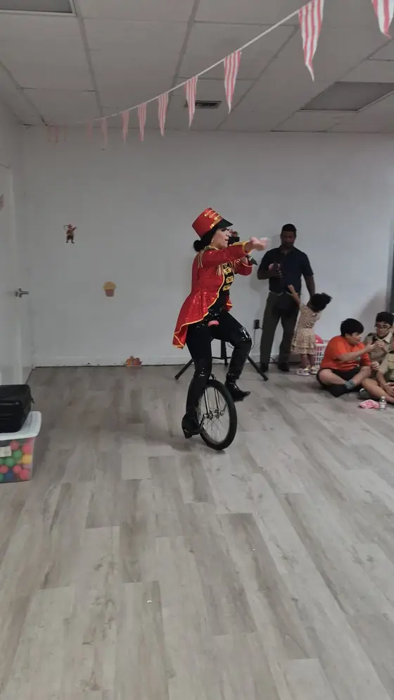 A person in a red ringmaster jacket and top hat rides a unicycle at an indoor circus-themed pack event, with Cub Scouts watching.