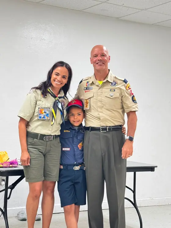Mr. Bob, Cubmaster and Webelos Den Leader, stands in a tan uniform beside Ms. Ceci, Bears Den Leader, with Cub Scouts at an indoor pack meeting.
