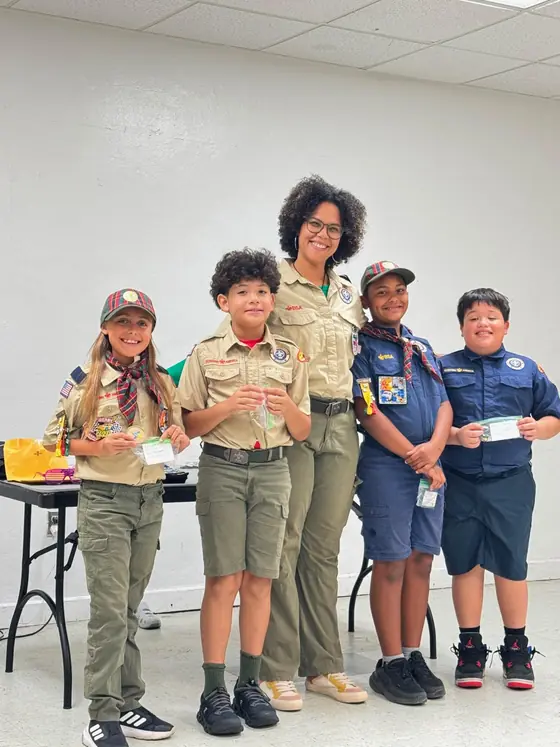Ms. Ivette, Assistant Cubmaster and Arrow of Light Den Leader, stands with Cub Scouts at an indoor pack meeting.