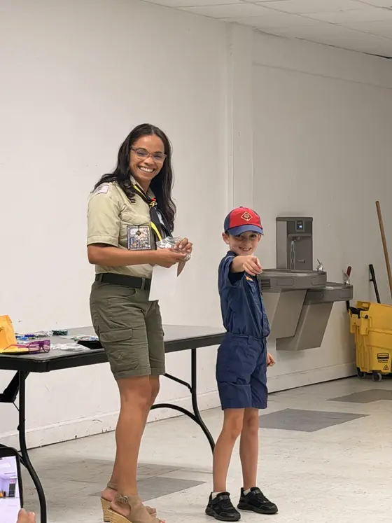 Ms. Ceci, Bears Den Leader, stands with a Cub Scout at an indoor pack meeting.