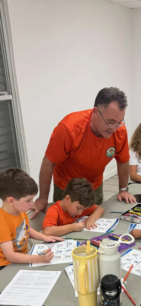 Mr. Rudy, Tigers and Lions Den Leader, wearing glasses and an orange pack shirt, leans over a table helping Cub Scouts with a worksheet.